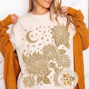 Pode incluir: Camiseta creme com um design celestial dourado, apresentando uma lua crescente, estrelas e elementos florais. A camiseta tem gola redonda e mangas curtas. Design original da Coco Bear.
