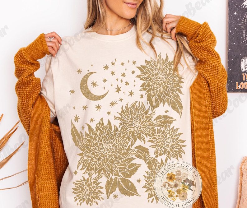 Pode incluir: Camiseta creme com um design celestial dourado, apresentando uma lua crescente, estrelas e elementos florais. A camiseta tem gola redonda e mangas curtas. Design original da Coco Bear.