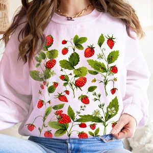 Puede incluir: Una sudadera rosa claro con un dise&ntilde;o de acuarela de fresas, hojas verdes y flores blancas. Las fresas son rojas y las hojas son verdes. El dise&ntilde;o se extiende por la parte delantera de la sudadera.