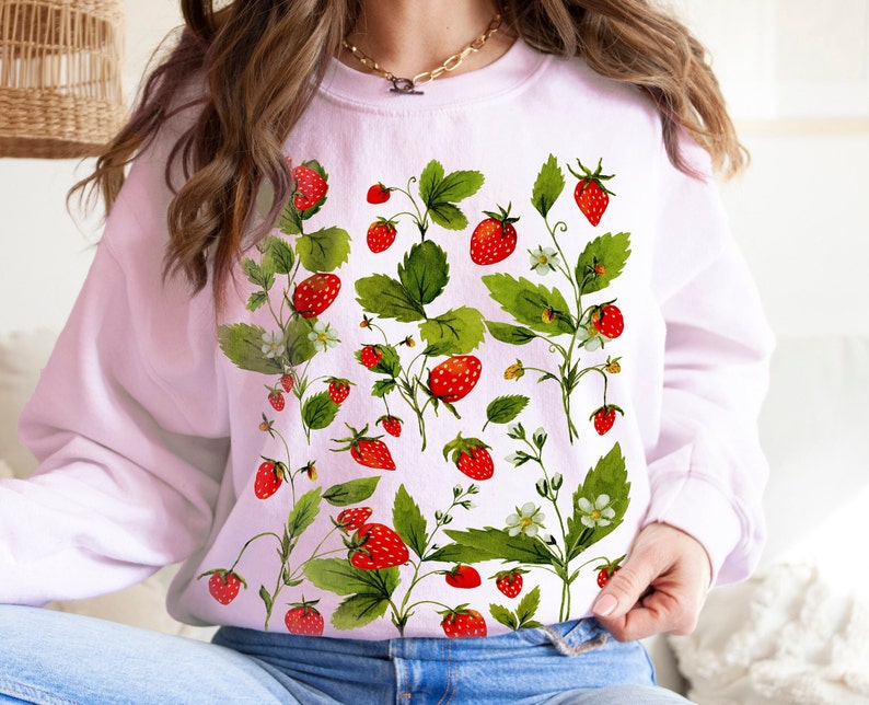 Puede incluir: Una sudadera rosa claro con un dise&ntilde;o de acuarela de fresas, hojas verdes y flores blancas. Las fresas son rojas y las hojas son verdes. El dise&ntilde;o se extiende por la parte delantera de la sudadera.