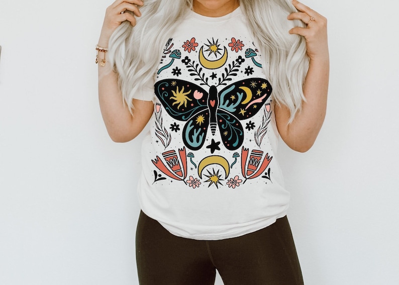 May include: A white t-shirt featuring a colourful butterfly design. The butterfly is surrounded by flowers, stars and moons. The design is in a folk art style.