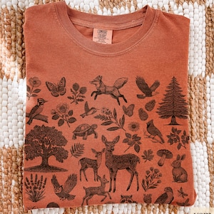 May include: Rust-colored crew neck t-shirt with a detailed woodland animal and plant design. The graphic includes a fox, deer, owl, tree, and other forest elements. The shirt has a Comfort Colors tag.