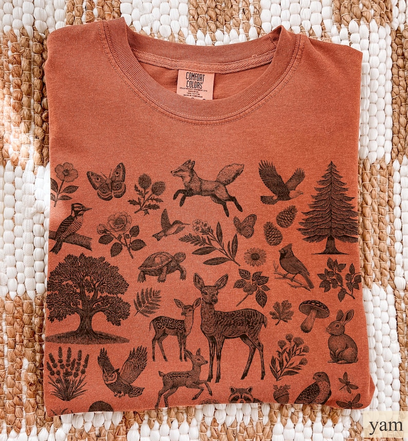 May include: Rust-colored crew neck t-shirt with a detailed woodland animal and plant design. The graphic includes a fox, deer, owl, tree, and other forest elements. The shirt has a Comfort Colors tag.