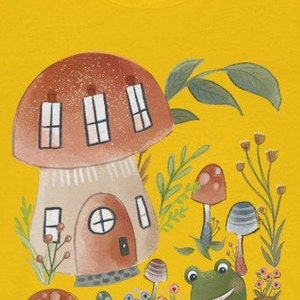 K&ouml;nnte beinhalten: Ein gelbes T-Shirt mit einer Cartoon-Illustration eines Pilzhauses, eines Frosches und anderer Pilze. Das Pilzhaus hat ein braunes Dach und einen beigefarbenen K&ouml;rper mit Fenstern und einer T&uuml;r. Der Frosch ist gr&uuml;n mit einem gro&szlig;en L&auml;cheln und sitzt auf dem Boden. Die Pilze sind rot und braun mit wei&szlig;en Flecken.