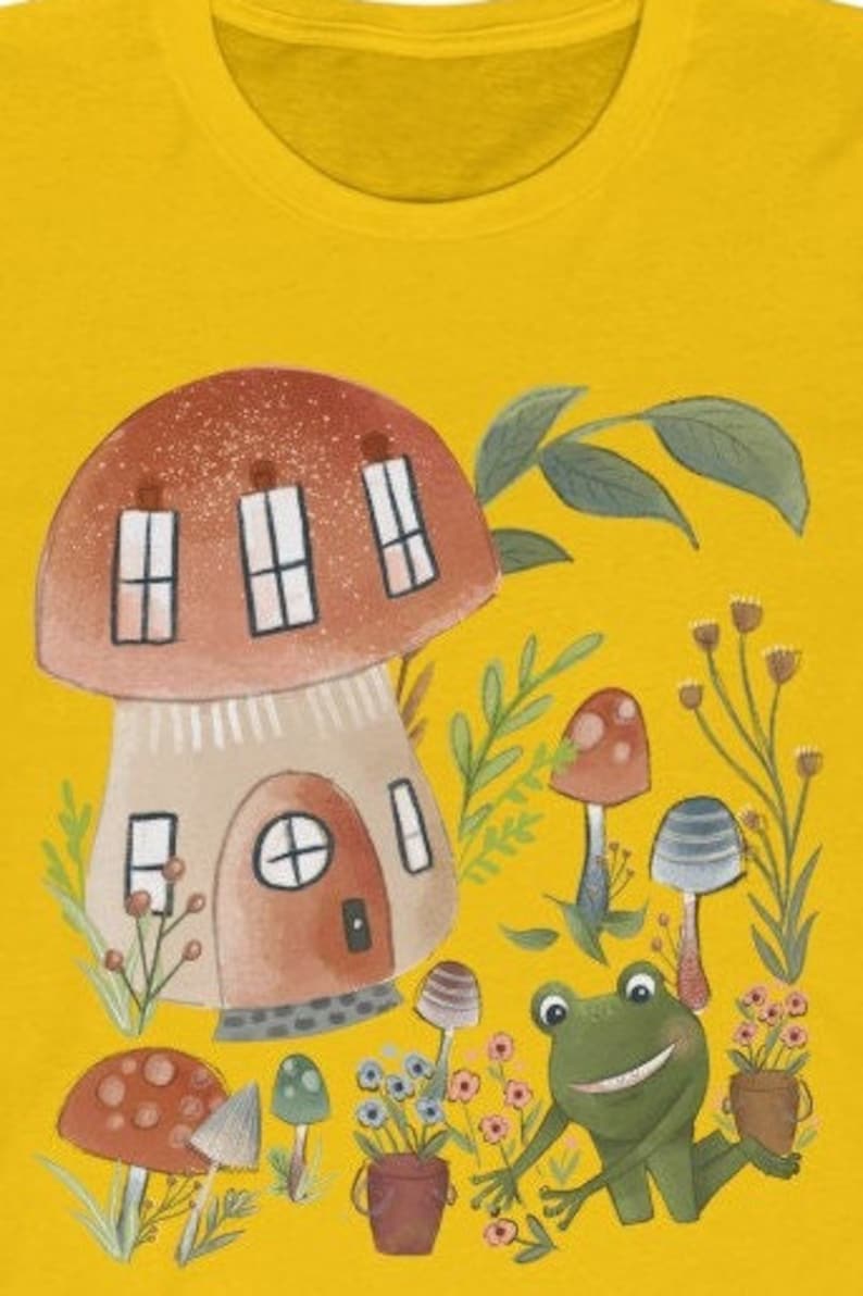 K&ouml;nnte beinhalten: Ein gelbes T-Shirt mit einer Cartoon-Illustration eines Pilzhauses, eines Frosches und anderer Pilze. Das Pilzhaus hat ein braunes Dach und einen beigefarbenen K&ouml;rper mit Fenstern und einer T&uuml;r. Der Frosch ist gr&uuml;n mit einem gro&szlig;en L&auml;cheln und sitzt auf dem Boden. Die Pilze sind rot und braun mit wei&szlig;en Flecken.