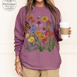 May include: A berry-colored long-sleeve sweatshirt featuring a colorful graphic print of various wildflowers, a monarch butterfly, and a small bee. The design includes red, yellow, purple, and orange floral elements on the front of the garment.
