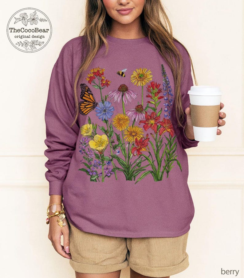 May include: A berry-colored long-sleeve sweatshirt featuring a colorful graphic print of various wildflowers, a monarch butterfly, and a small bee. The design includes red, yellow, purple, and orange floral elements on the front of the garment.