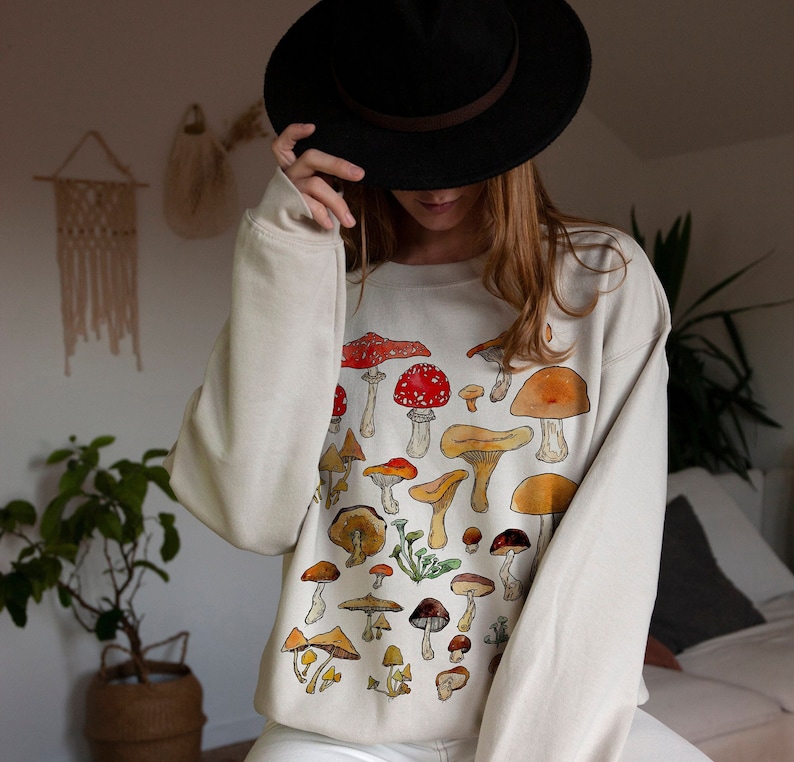 May include: A cream-colored sweatshirt with a mushroom print. The mushrooms are in various shades of brown, red, and yellow. The sweatshirt has long sleeves and a crew neck.