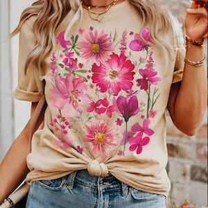 May include: A beige t-shirt adorned with a colourful floral print in pink and magenta hues. The flowers are clustered across the front. The shirt is knotted at the waist and worn with denim shorts.