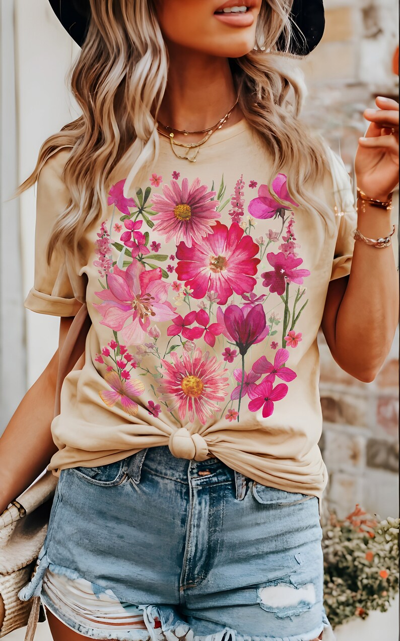 May include: A beige t-shirt adorned with a colourful floral print in pink and magenta hues. The flowers are clustered across the front. The shirt is knotted at the waist and worn with denim shorts.