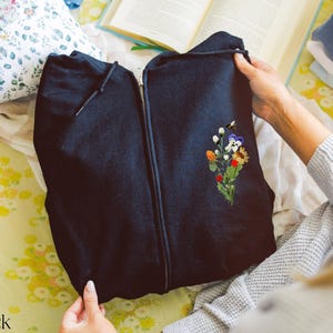 May include: Black zip-up hoodie with a floral embroidery design featuring a bee, flowers, and leaves.