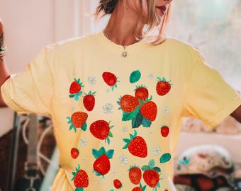 Puede incluir: Una camiseta amarilla con estampado de fresas. Las fresas son rojas con hojas verdes y están dispersas por la camiseta.