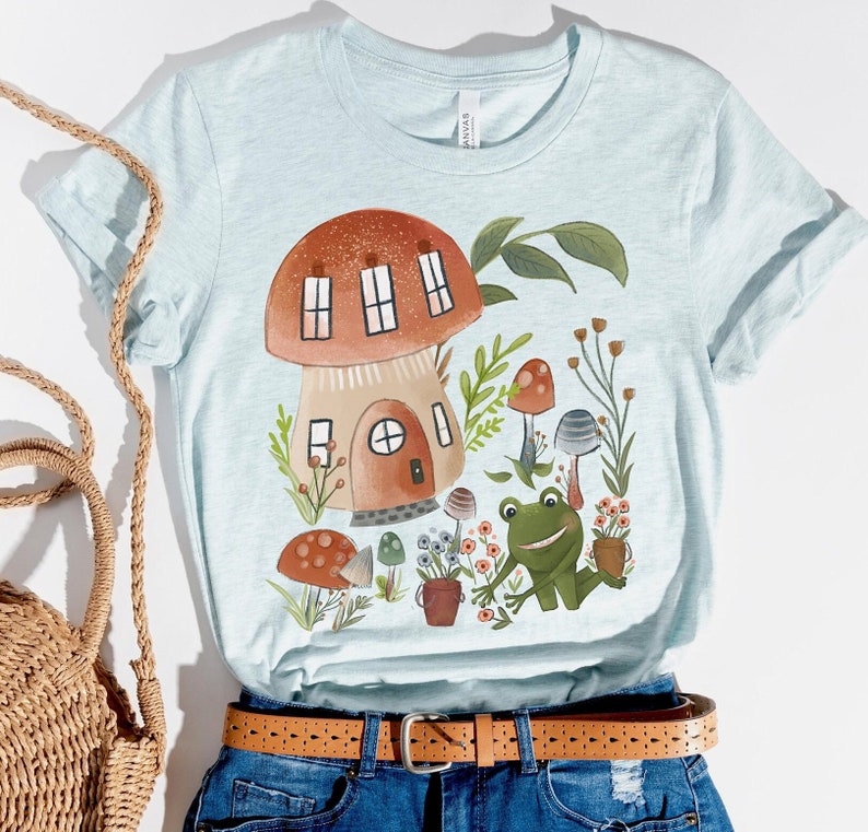 K&ouml;nnte beinhalten: Ein hellblaues T-Shirt mit einer fantasievollen Illustration eines Pilzhauses, eines Frosches und anderer Waldwesen. Das Design zeigt einen braunen Pilz mit einer wei&szlig;en T&uuml;r und Fenstern, einen gr&uuml;nen Frosch mit wei&szlig;em Bauch und verschiedene Pilze und Pflanzen in Gr&uuml;n-, Braun- und Rott&ouml;nen.