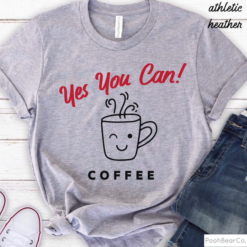 Może przedstawiać: Szara koszulka athletic heather z czerwonym napisem "Yes You Can!" nad grafiką mrugającego kubka z kawą. Słowo "COFFEE" jest wydrukowane na dole czarnymi literami. Koszulka jest zestawiona z niebieskimi dżinsami.