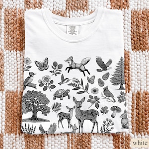 May include: White t-shirt with a black and white nature-themed graphic. The design includes illustrations of a fox, eagle, deer, butterflies, birds, and various plants. The shirt is folded and displayed on a textured surface.
