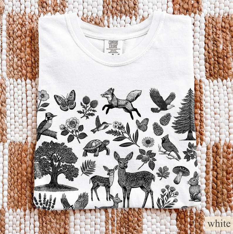 May include: White t-shirt with a black and white nature-themed graphic. The design includes illustrations of a fox, eagle, deer, butterflies, birds, and various plants. The shirt is folded and displayed on a textured surface.