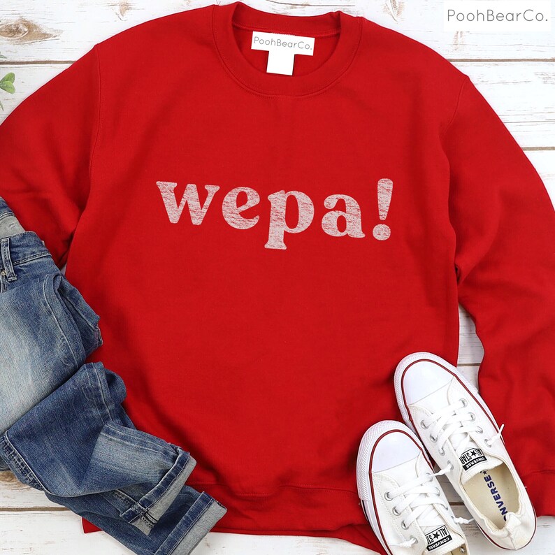 May include: Red sweatshirt with the word "wepa!" printed in white on the front. The sweatshirt is laying on a white wooden surface with a pair of white sneakers and blue jeans.