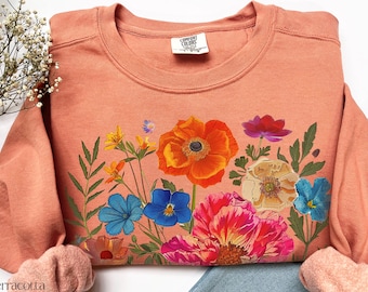 Op de afbeelding: Een terracotta kleurige sweatshirt met een levendig bloemmotief. Het ontwerp bevat kleurrijke bloemen in tinten oranje, blauw, roze en paars, met groene bladeren. De sweatshirt heeft een ronde hals en lange mouwen.