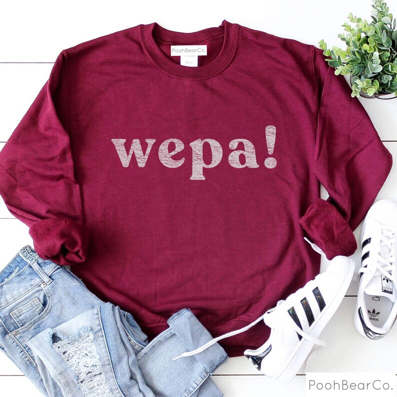 May include: Burgundy sweatshirt with the word "wepa!" printed in white on the front. The sweatshirt is laying on a white surface with a pair of white sneakers and blue jeans.