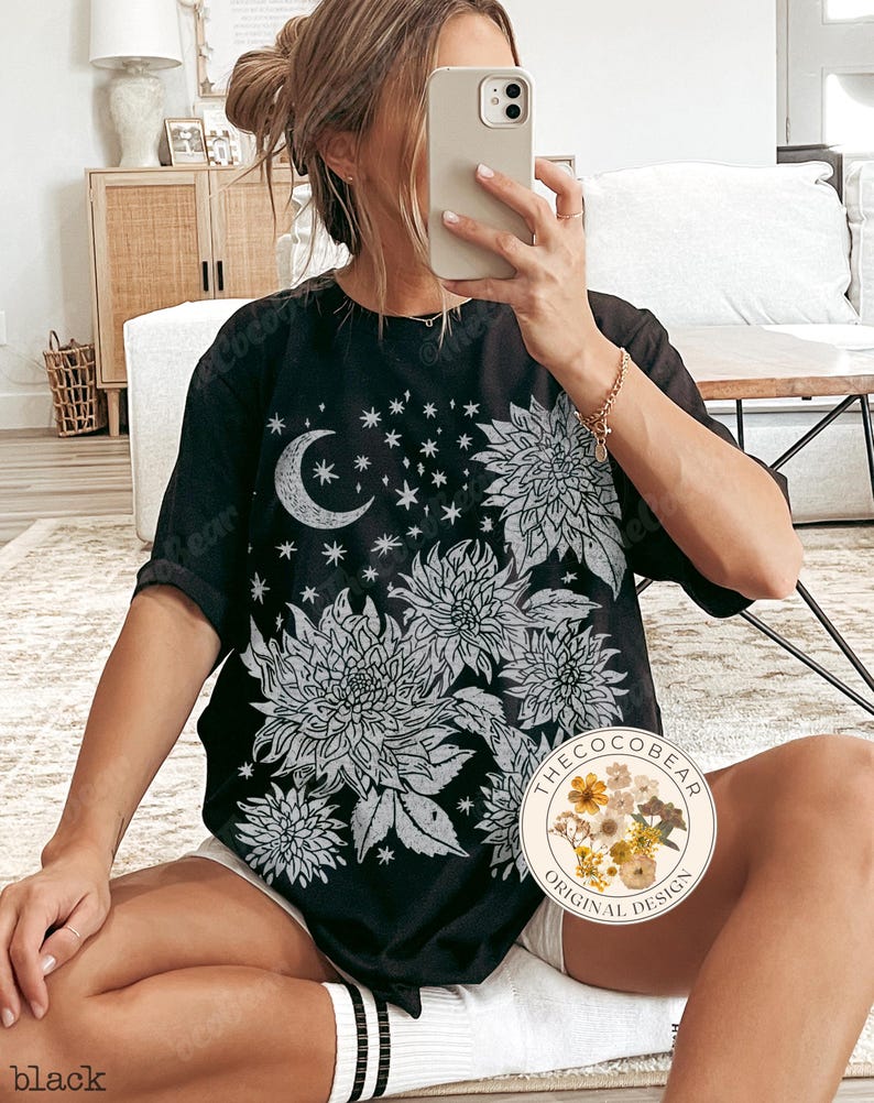 Pode incluir: Camiseta preta com um design floral e celestial branco. O design inclui uma lua crescente, estrelas e grandes ilustra&ccedil;&otilde;es de flores. A camiseta tem mangas curtas e um corte folgado. O texto "black" &eacute; vis&iacute;vel na parte inferior esquerda.