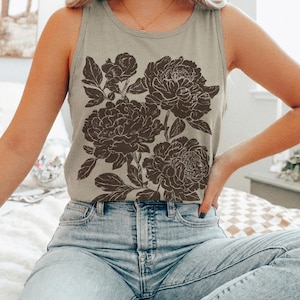 May include: A light olive green sleeveless tank top featuring a dark brown floral graphic print on the front. The design displays detailed illustrations of blooming peonies with leaves and stems. The garment has a round neckline and is paired with light blue denim jeans. The word sandstone is printed in the bottom right corner.