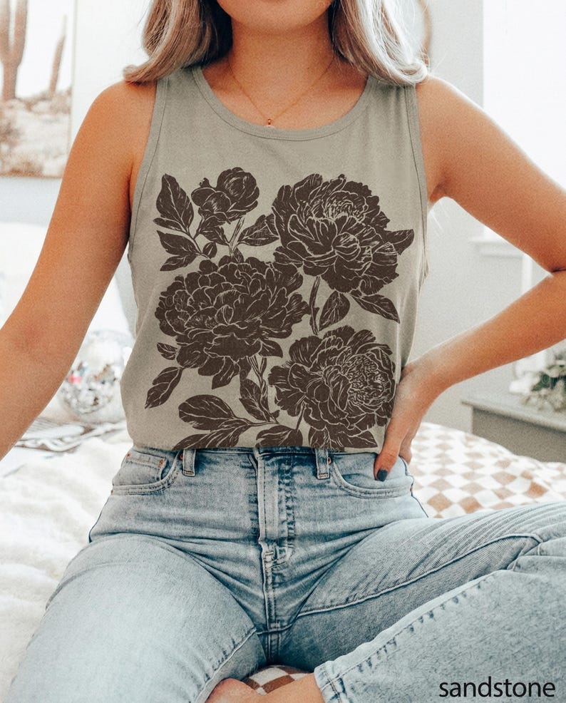 May include: A light olive green sleeveless tank top featuring a dark brown floral graphic print on the front. The design displays detailed illustrations of blooming peonies with leaves and stems. The garment has a round neckline and is paired with light blue denim jeans. The word sandstone is printed in the bottom right corner.