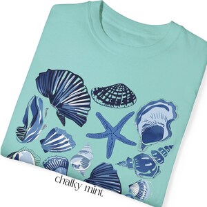 Puede incluir: Una camiseta azul turquesa con un dise&ntilde;o de varias conchas marinas azules y blancas y una estrella de mar. El texto "chalky mint" est&aacute; impreso en la parte inferior de la camiseta.
