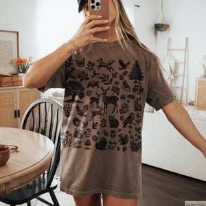 May include: Espresso-colored t-shirt featuring a detailed black illustration of woodland creatures and plants. The design includes deer, birds, foxes, and trees. The shirt has a relaxed fit and short sleeves, perfect for casual wear.