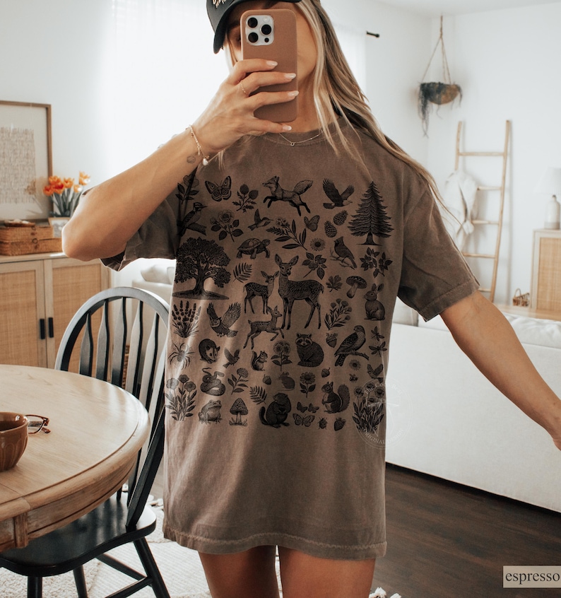 May include: Espresso-colored t-shirt featuring a detailed black illustration of woodland creatures and plants. The design includes deer, birds, foxes, and trees. The shirt has a relaxed fit and short sleeves, perfect for casual wear.