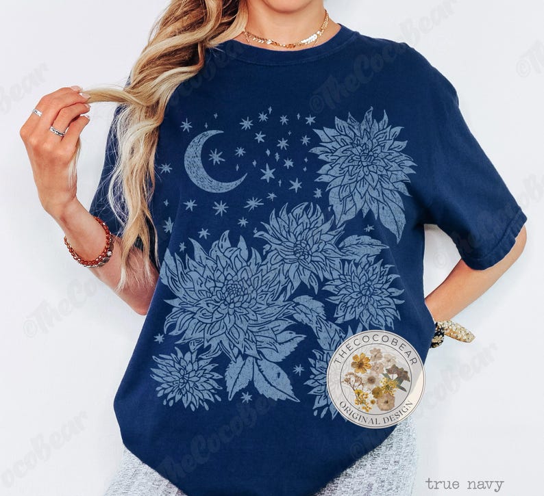 Pode incluir: Uma camiseta azul marinho com um design floral e celestial. O design inclui uma lua crescente, estrelas e v&aacute;rias flores grandes e detalhadas em um tom de azul mais claro. A camiseta tem decote redondo e mangas curtas.