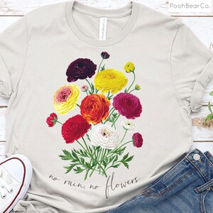 May include: A light gray t-shirt with a floral design featuring a bouquet of colorful ranunculus flowers in shades of red, yellow, white, and burgundy. The text "no rain, no flowers" is printed below the flowers.
