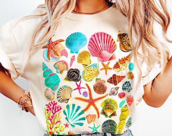 Op de afbeelding: Ivoor kleurige sweatshirt met een levendige verzameling schelpen en zeesterren in verschillende kleuren. Het ontwerp bevat roze, blauwe, gele en groene schelpen, wat een zomerse, strandthema esthetiek creëert. De tekst "TheCocoBear" is zichtbaar.
