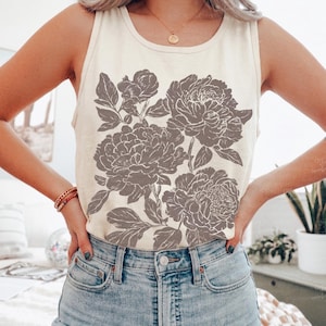 May include: An ivory sleeveless tank top featuring a large, detailed graphic print of blooming peonies in a muted brown tone. The shirt has a classic round neckline and is paired with light blue denim jeans, creating a casual look.