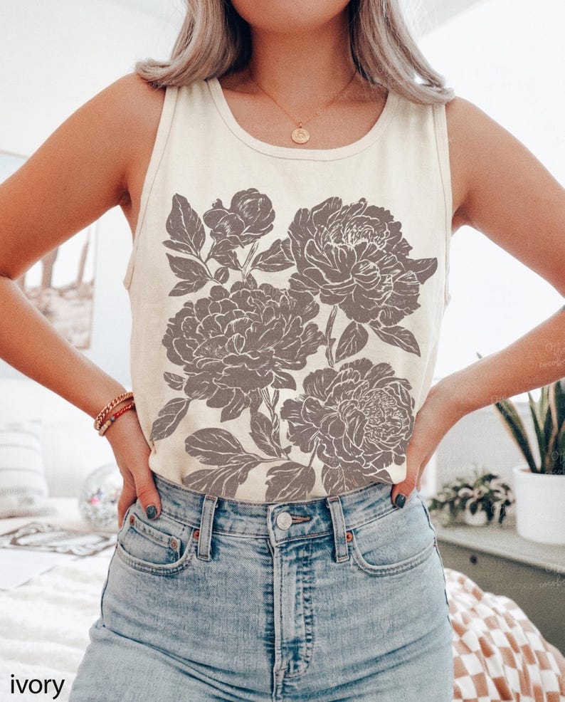 May include: An ivory sleeveless tank top featuring a large, detailed graphic print of blooming peonies in a muted brown tone. The shirt has a classic round neckline and is paired with light blue denim jeans, creating a casual look.