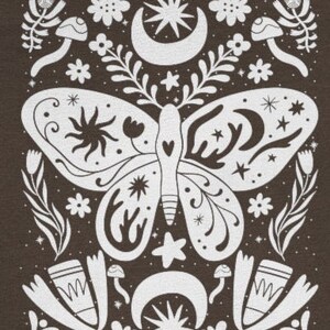 Puede incluir: Un dise&ntilde;o gr&aacute;fico blanco sobre un fondo marr&oacute;n presenta una mariposa estilizada con elementos florales y celestiales. La mariposa tiene un cuerpo en forma de coraz&oacute;n y alas intrincadas con estrellas, lunas y soles. El dise&ntilde;o est&aacute; rodeado de flores, hongos y lunas crecientes.