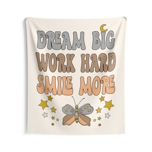 May include: A beige tapestry with the text "Dream Big Work Hard Smile More" in a retro font. The text is in shades of gray, brown, and peach. A butterfly with gray, brown, and peach wings is centered below the text. The butterfly is surrounded by gold and white stars.