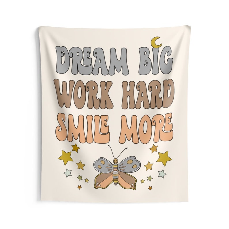 May include: A beige tapestry with the text "Dream Big Work Hard Smile More" in a retro font. The text is in shades of gray, brown, and peach. A butterfly with gray, brown, and peach wings is centered below the text. The butterfly is surrounded by gold and white stars.