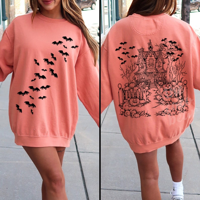Op de afbeelding: Een koraalkleurig sweatshirt met een zwarte afbeelding van vleermuizen op de voorkant en een zwarte afbeelding van een spookhuis met pompoenen op de achterkant.