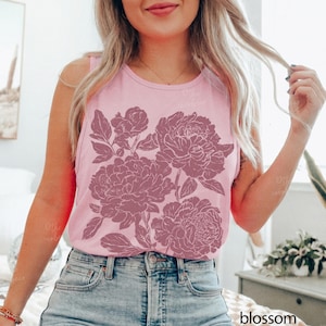 May include: A light pink sleeveless tank top featuring a large, dark pink floral graphic print of peonies on the front. The garment has a round neckline and is paired with blue denim jeans. The word blossom is printed in the bottom right corner.