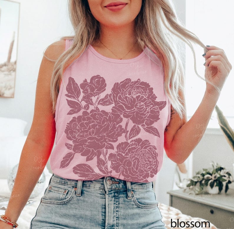 May include: A light pink sleeveless tank top featuring a large, dark pink floral graphic print of peonies on the front. The garment has a round neckline and is paired with blue denim jeans. The word blossom is printed in the bottom right corner.