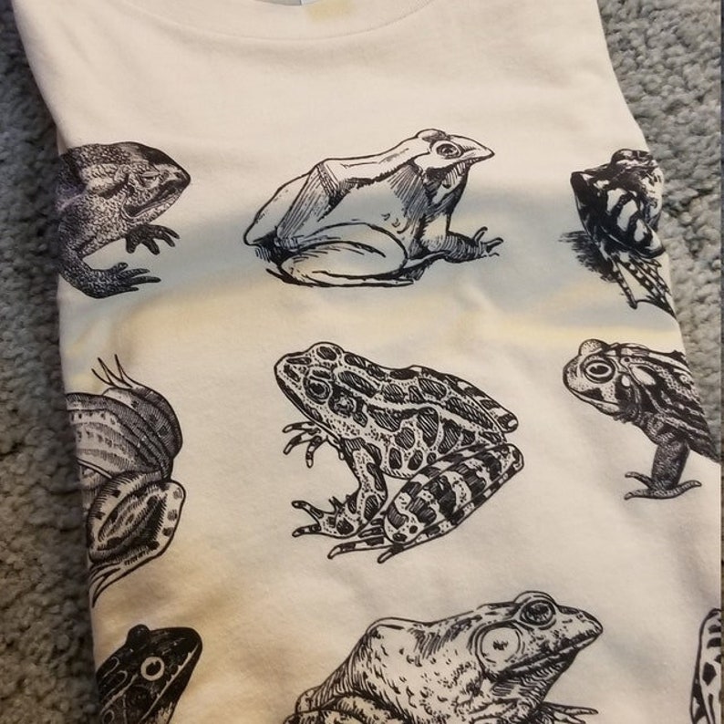Puede incluir: Camiseta de color crema con un patr&oacute;n repetido de ilustraciones detalladas de ranas en blanco y negro. Las ranas var&iacute;an en tama&ntilde;o y pose, creando un dise&ntilde;o caprichoso. La tela de la camiseta parece suave y c&oacute;moda.