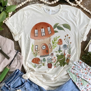 K&ouml;nnte beinhalten: Ein cremefarbenes T-Shirt mit einer Illustration eines Pilzhauses. Das Haus hat ein rotes Dach und braune W&auml;nde. Die Illustration enth&auml;lt gr&uuml;ne Bl&auml;tter, Blumen und einen Frosch.