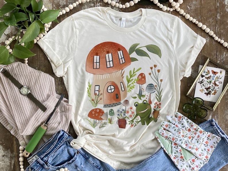 K&ouml;nnte beinhalten: Ein cremefarbenes T-Shirt mit einer Illustration eines Pilzhauses. Das Haus hat ein rotes Dach und braune W&auml;nde. Die Illustration enth&auml;lt gr&uuml;ne Bl&auml;tter, Blumen und einen Frosch.