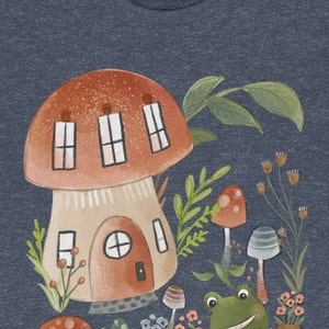 K&ouml;nnte beinhalten: Ein blaues T-Shirt mit einer fantasievollen Illustration eines Pilzhauses, eines Frosches und anderer Pilze. Das Pilzhaus hat ein rotes Dach und braune W&auml;nde mit Fenstern. Der Frosch ist gr&uuml;n mit einem breiten L&auml;cheln. Die Pilze sind rot und braun mit wei&szlig;en Flecken.