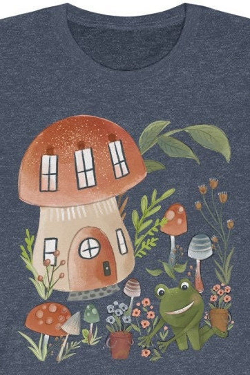 K&ouml;nnte beinhalten: Ein blaues T-Shirt mit einer fantasievollen Illustration eines Pilzhauses, eines Frosches und anderer Pilze. Das Pilzhaus hat ein rotes Dach und braune W&auml;nde mit Fenstern. Der Frosch ist gr&uuml;n mit einem breiten L&auml;cheln. Die Pilze sind rot und braun mit wei&szlig;en Flecken.