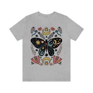 May include: A grey t-shirt featuring a colourful butterfly design. The butterfly is surrounded by stars, moons and flowers. The design is in a folk art style.