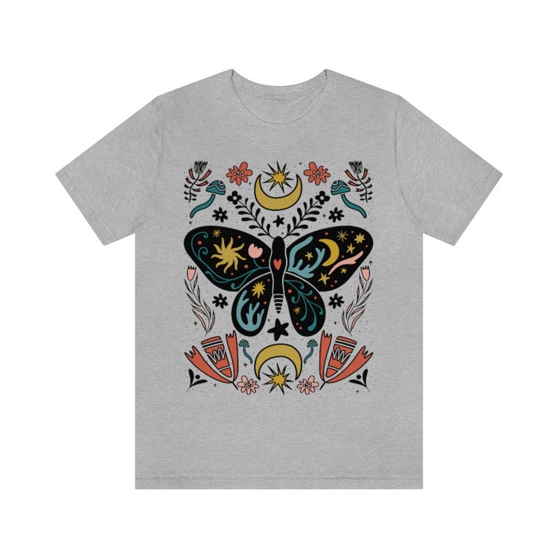 May include: A grey t-shirt featuring a colourful butterfly design. The butterfly is surrounded by stars, moons and flowers. The design is in a folk art style.