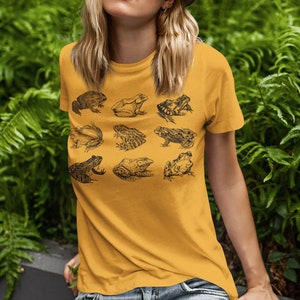Puede incluir: Camiseta de color amarillo mostaza con una cuadr&iacute;cula de nueve ilustraciones detalladas de ranas negras. Las ranas, de varios tama&ntilde;os y poses, muestran diferentes especies. La camiseta, con cuello redondo y mangas cortas, es ideal para un look casual.