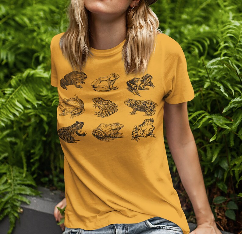 Puede incluir: Camiseta de color amarillo mostaza con una cuadr&iacute;cula de nueve ilustraciones detalladas de ranas negras. Las ranas, de varios tama&ntilde;os y poses, muestran diferentes especies. La camiseta, con cuello redondo y mangas cortas, es ideal para un look casual.