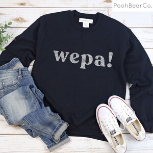 May include: A black sweatshirt with the word "wepa!" printed in white on the front. The sweatshirt is being modeled with blue jeans and white sneakers.